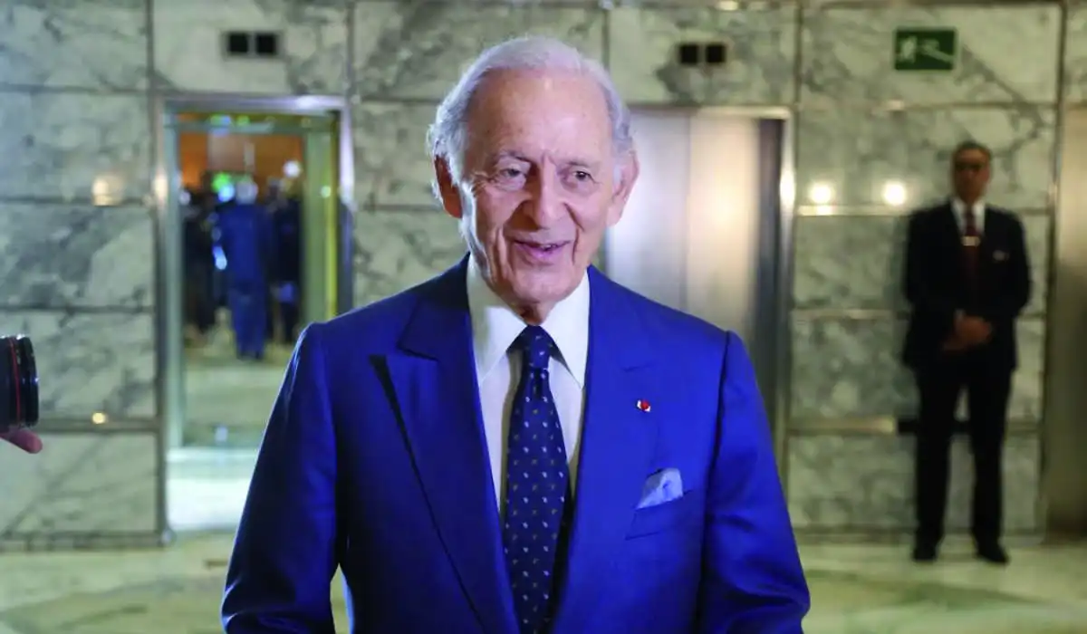 An older man in a stylish blue suit and matching tie, carrying a pocket square and a boutonniere decoration, standing in a well-lit marble hall, with security agents in the background near open elevators.